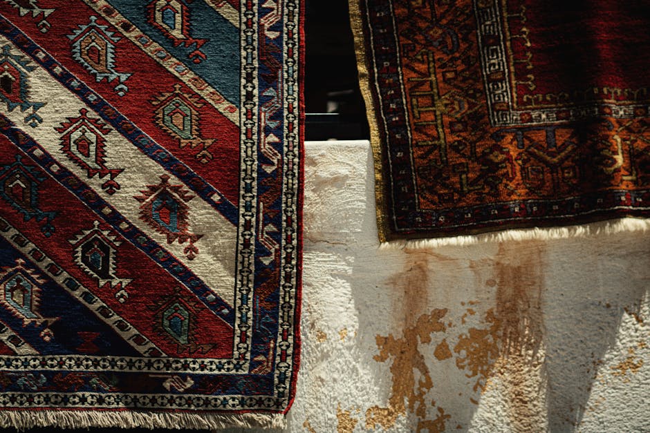 Close-up image of two Persian-style area rugs with intricate geometric and floral patterns in red, blue, beige, and orange tones, draped over a white textured surface. The top rug shows signs of stain