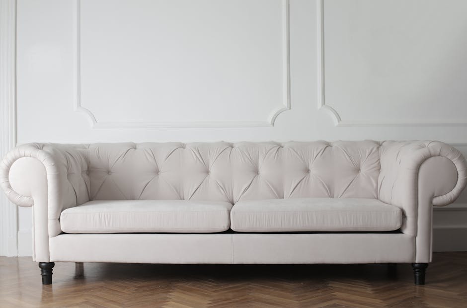 A close-up view of a person using a handheld upholstery cleaning machine on a fabric sofa cushion in a living room. The operator's hand, gripping the tool, is visible as the device applies deep cleani